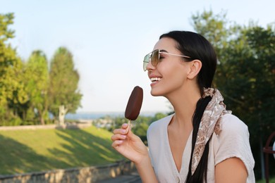 Photo of Beautiful young woman eating ice cream glazed in chocolate outdoors, space for text