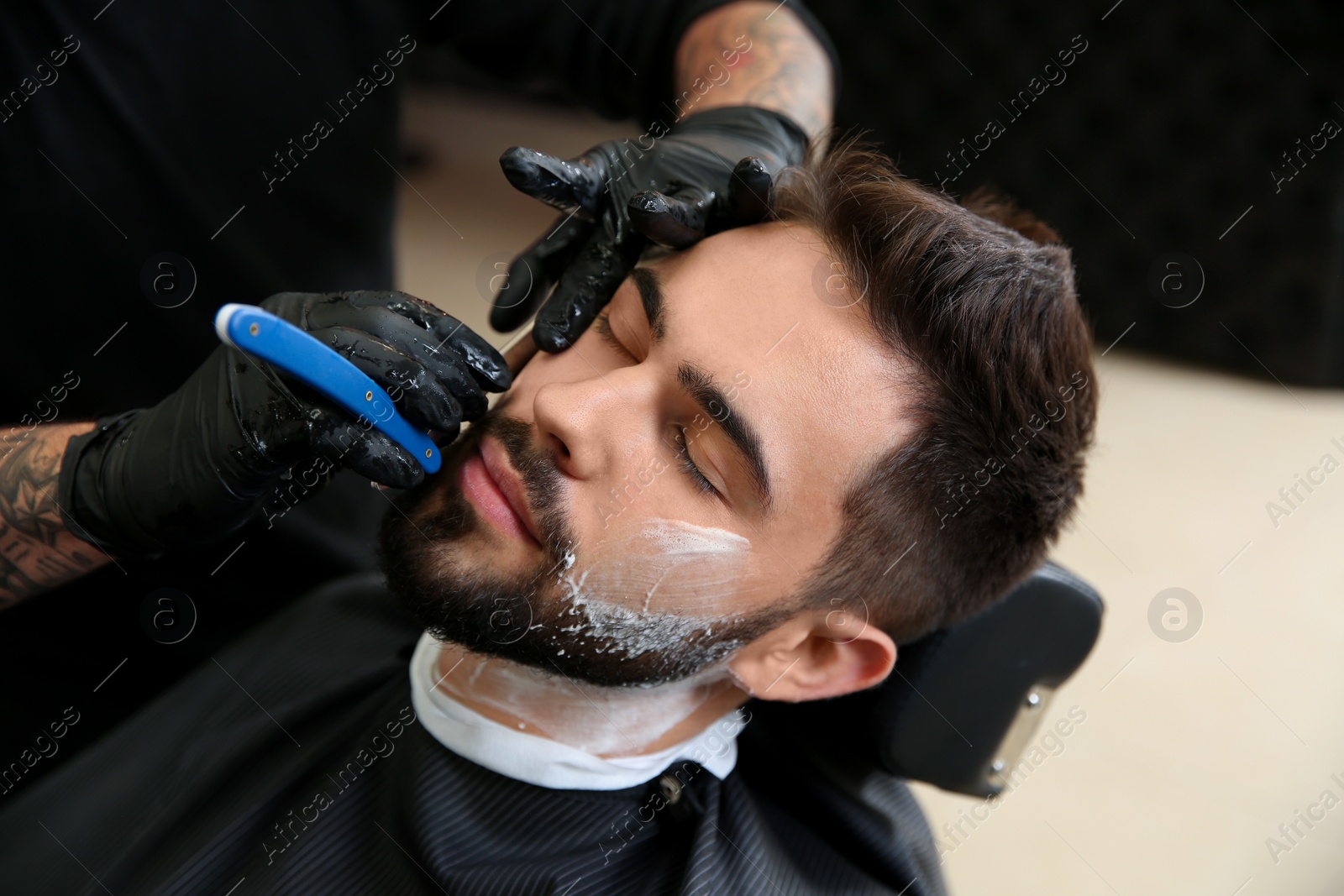 Photo of Professional hairdresser shaving client with straight razor in barbershop