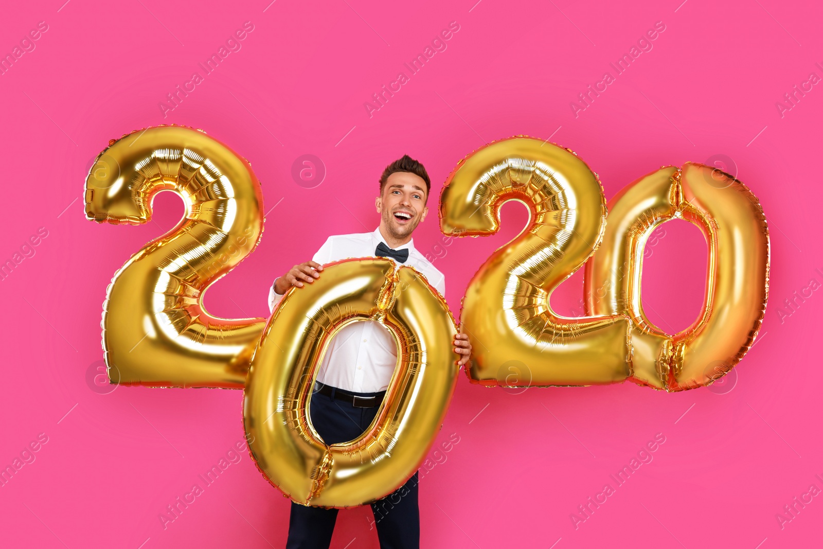 Photo of Happy young man with golden 2020 balloons on pink background. New Year celebration