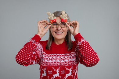 Photo of Happy senior woman in Christmas sweater and funny glasses on grey background
