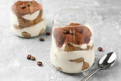 Photo of Delicious tiramisu in glasses, coffee beans and spoon on grey textured table