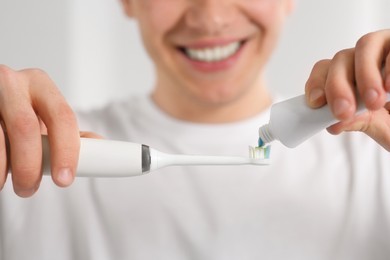 Man squeezing toothpaste from tube onto electric toothbrush on blurred background, closeup