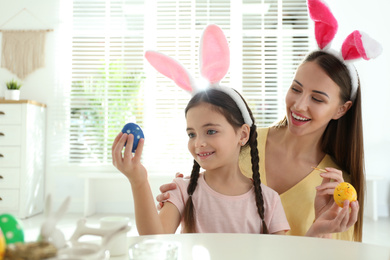 Photo of Happy mother and daughter with bunny ears headbands painting Easter eggs at home