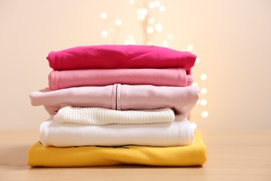 Photo of Stack of folded clothes on wooden table against beige background with blurred lights, space for text