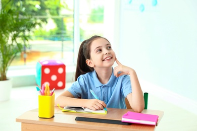 Pensive little girl doing assignment at desk in classroom. School stationery