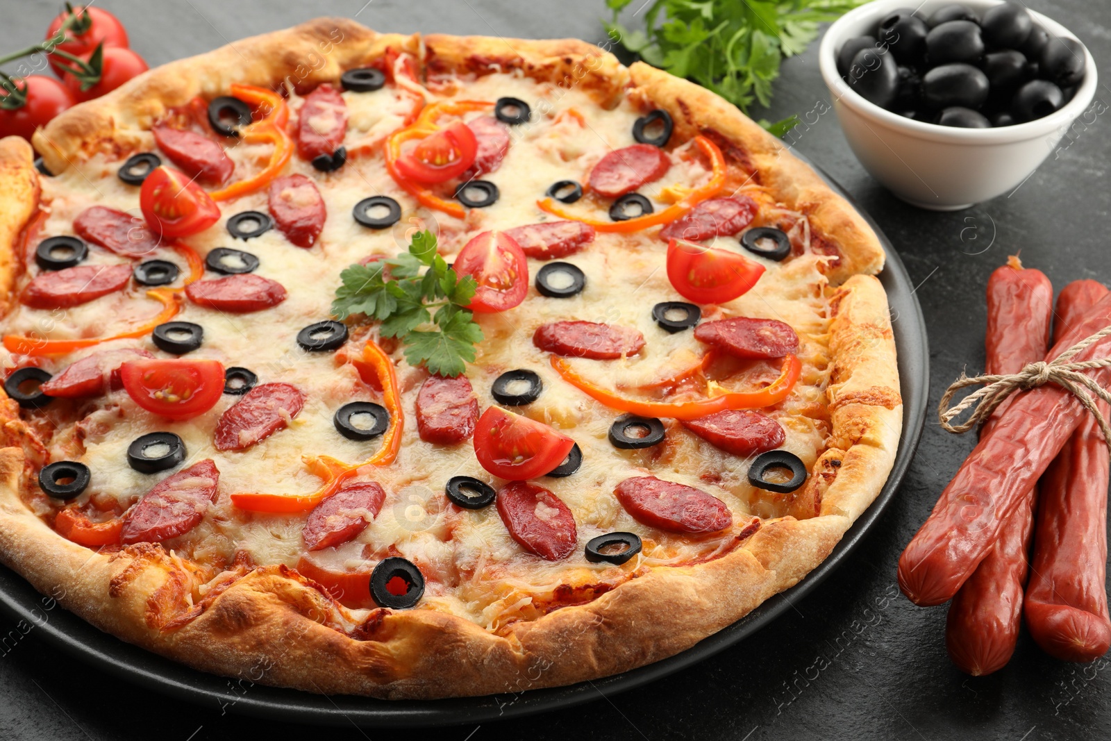 Photo of Tasty pizza with dry smoked sausages, olives, tomatoes, pepper and parsley on grey table