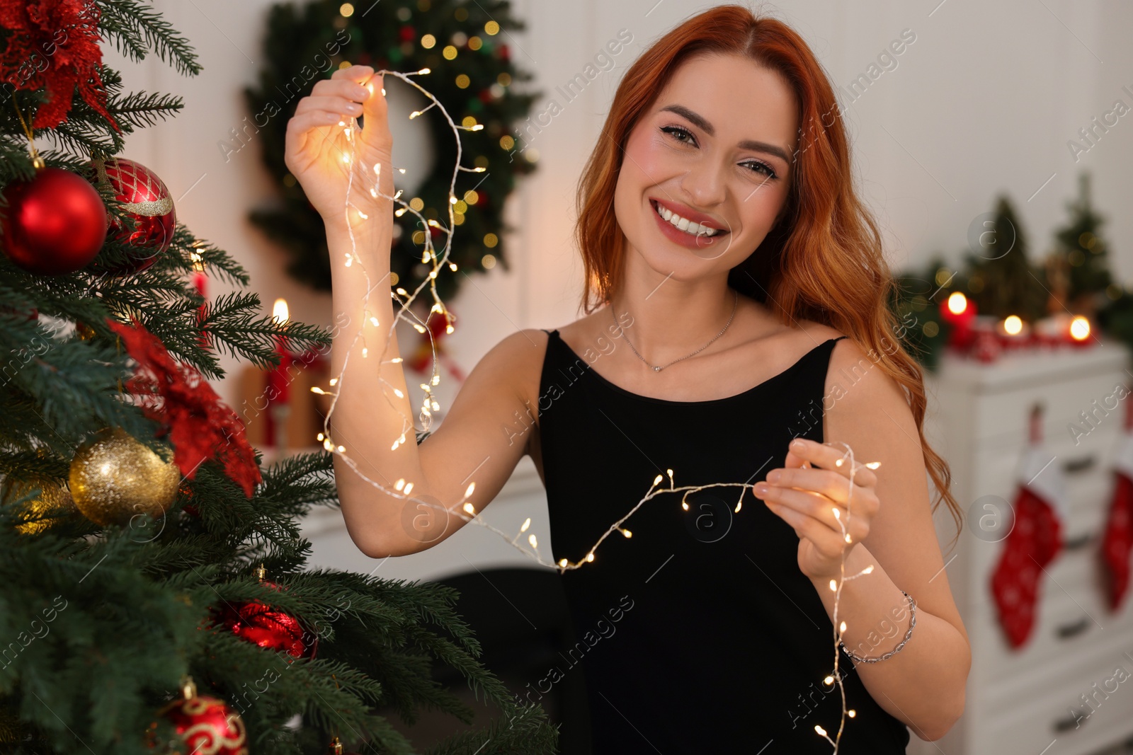 Photo of Happy young woman decorating Christmas tree with festive lights at home