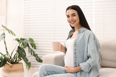 Happy pregnant woman with cup of drink on sofa at home