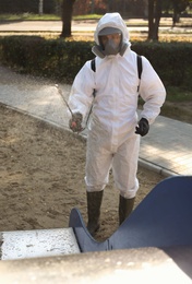 Photo of Man in hazmat suit spraying disinfectant onto slide at children's playground. Surface treatment during coronavirus pandemic