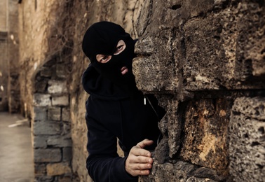 Photo of Man in mask behind stone wall watching someone outdoors. Criminal activity