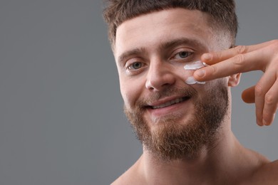 Photo of Handsome man applying moisturizing cream onto his face on grey background, space for text