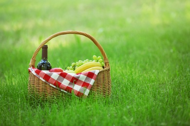 Photo of Wicker basket with blanket, wine and food on green grass in park, space for text. Summer picnic