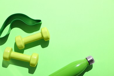 Photo of Dumbbells, fitness elastic band and water bottle on light green background, flat lay. Space for text