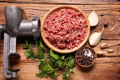 Photo of Manual meat grinder with beef mince, peppercorns, onion and parsley on wooden table, flat lay
