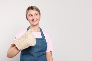 Photo of Beautiful young woman in denim apron and oven glove on light grey background. Space for text