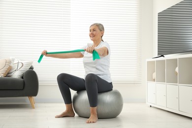 Photo of Senior woman doing exercise with elastic resistance band on fitness ball at home