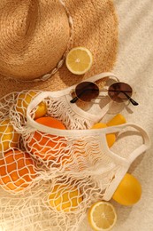Photo of String bag with sunglasses, straw hat and fruits on beige textured background, flat lay