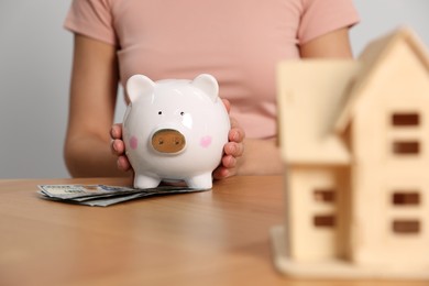 Photo of Woman holding piggy bank, house model and banknotes at wooden table, closeup