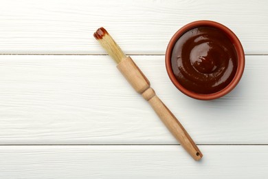 Photo of Tasty barbeque sauce in bowl and brush on white wooden table, top view. Space for text