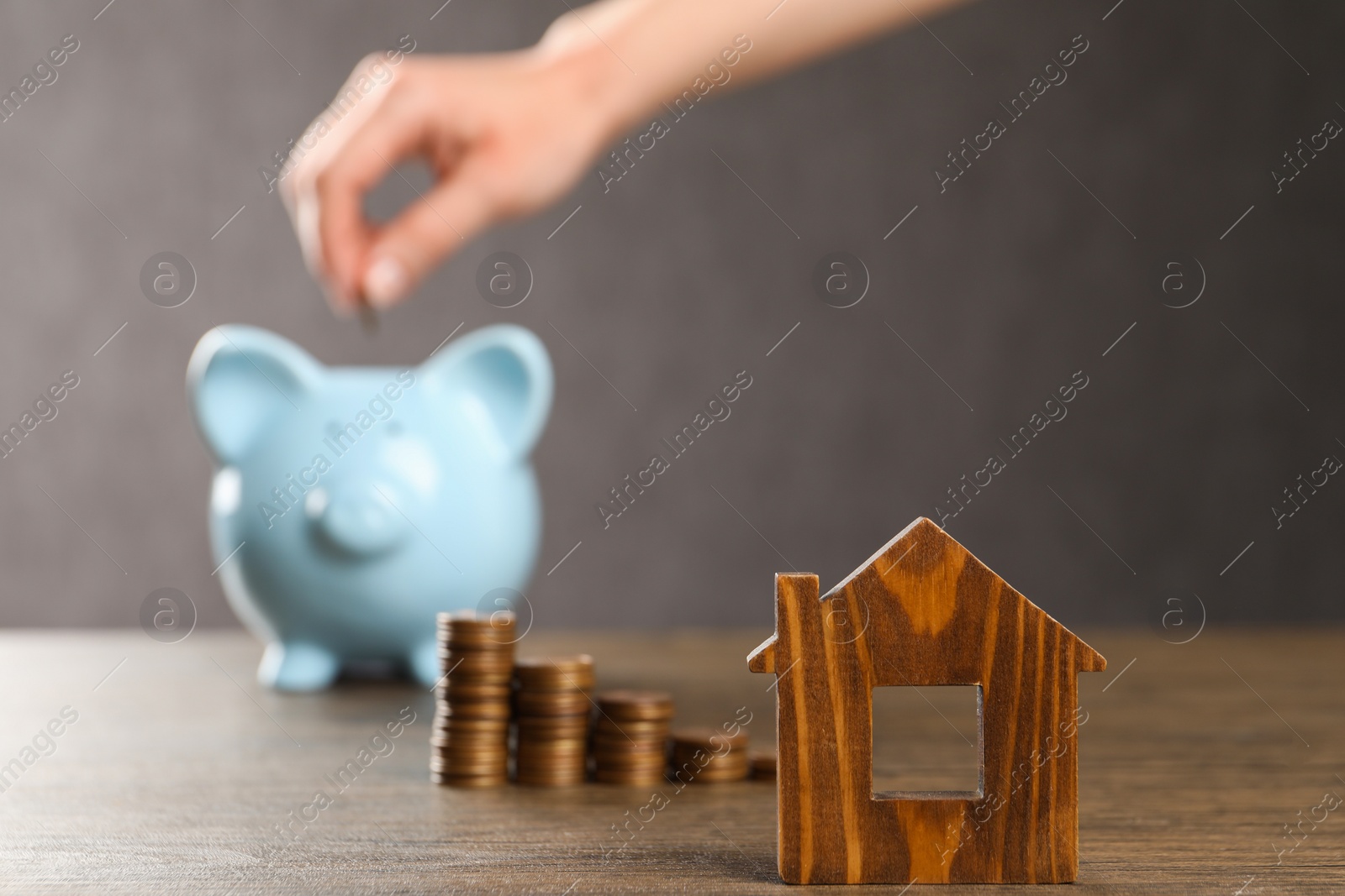 Photo of Woman putting coin into piggy bank at wooden table, focus on house model