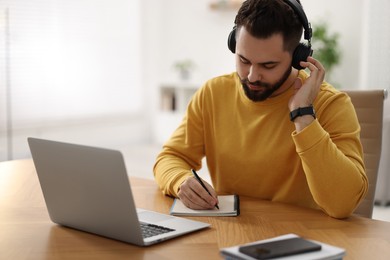 Photo of Young man in headphones writing down notes during webinar at table in room