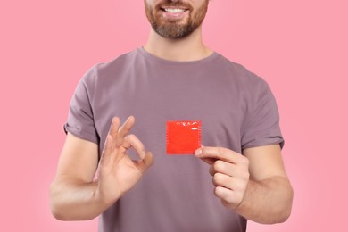 Photo of Man with condom showing ok gesture on pink background, closeup. Safe sex