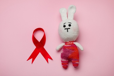 Photo of Cute knitted toy bunny and red ribbon on pink background, flat lay. AIDS disease awareness