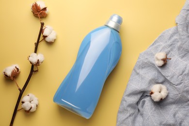 Photo of Bottle of fabric softener, cotton flowers and grey cloth on pale yellow background, flat lay