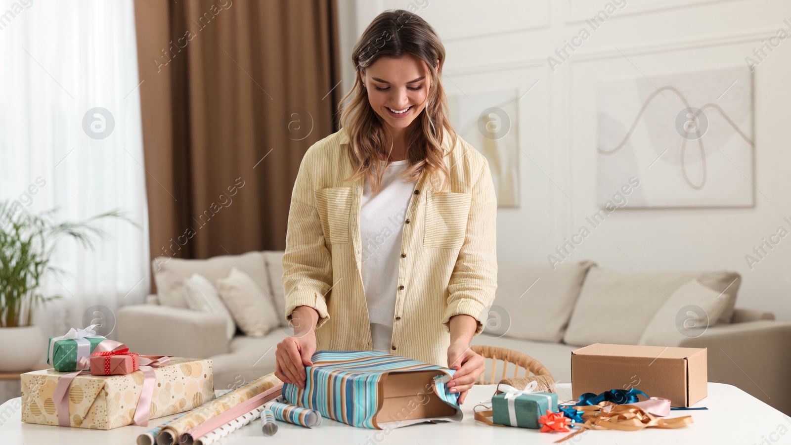 Photo of Beautiful young woman wrapping gift at table in living room