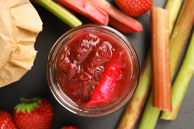 Photo of Jar of tasty rhubarb jam, fresh stems and strawberries on dark table, flat lay