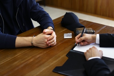 Photo of Police officer interrogating criminal in handcuffs at desk indoors