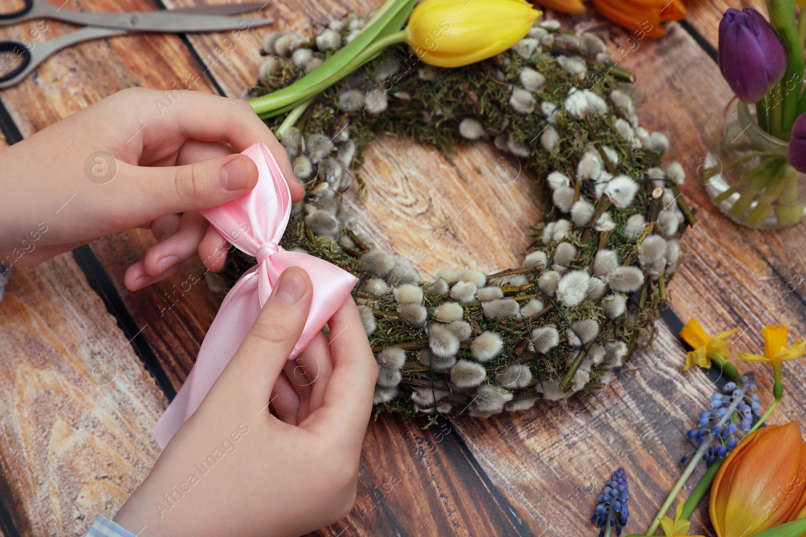 Photo of Woman decorating willow wreath with pink bow at wooden table, closeup