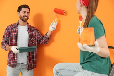 Photo of Man painting orange wall and woman holding can of dye with brush. Interior design