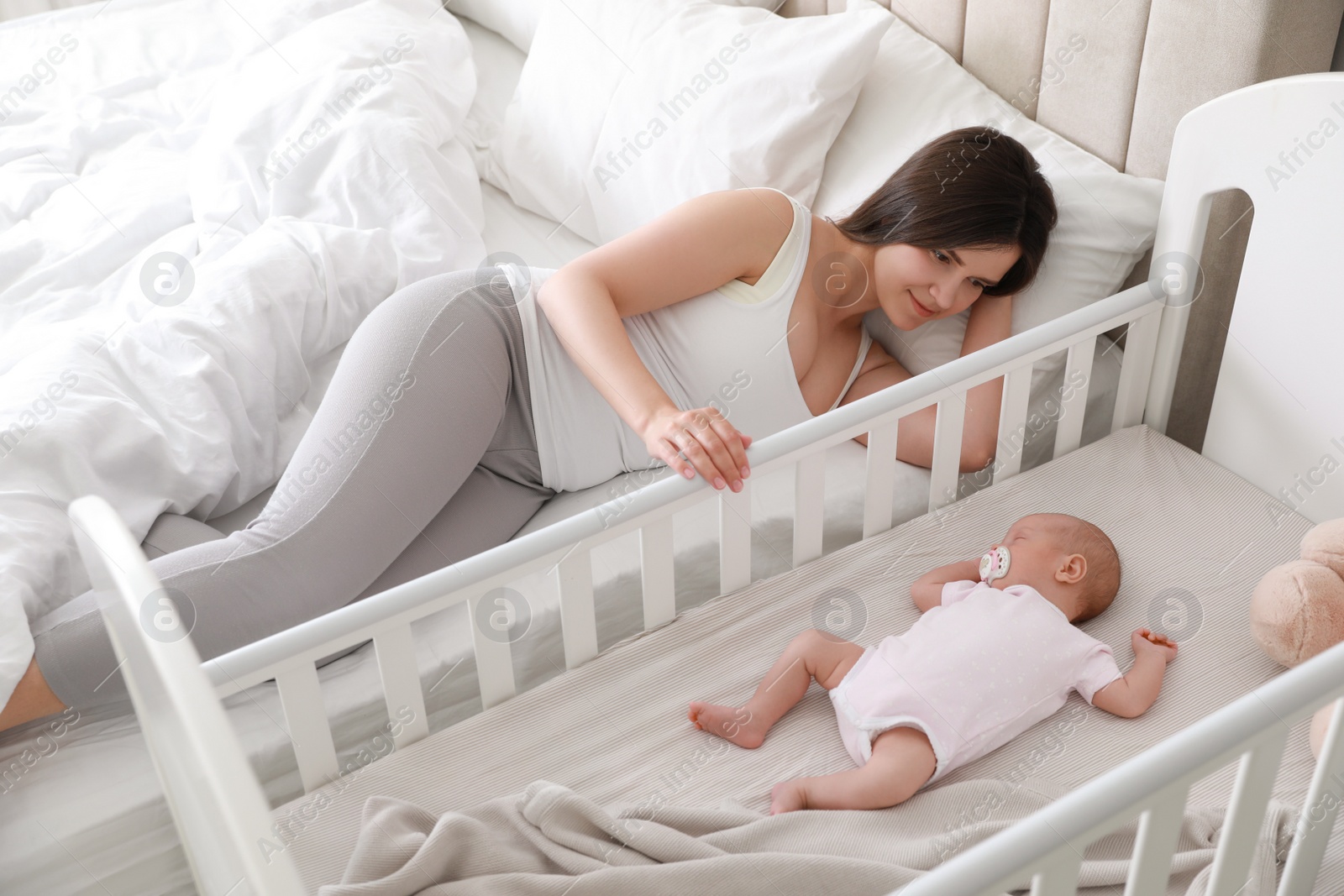 Photo of Happy young mother near crib with sleeping newborn baby in bedroom