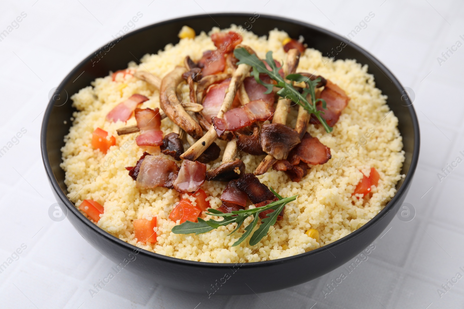 Photo of Tasty couscous with mushrooms and bacon in bowl on white tiled table, closeup