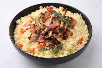 Tasty couscous with mushrooms and bacon in bowl on white tiled table, closeup