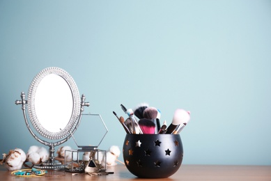 Holder with professional makeup brushes and mirror on table near color wall