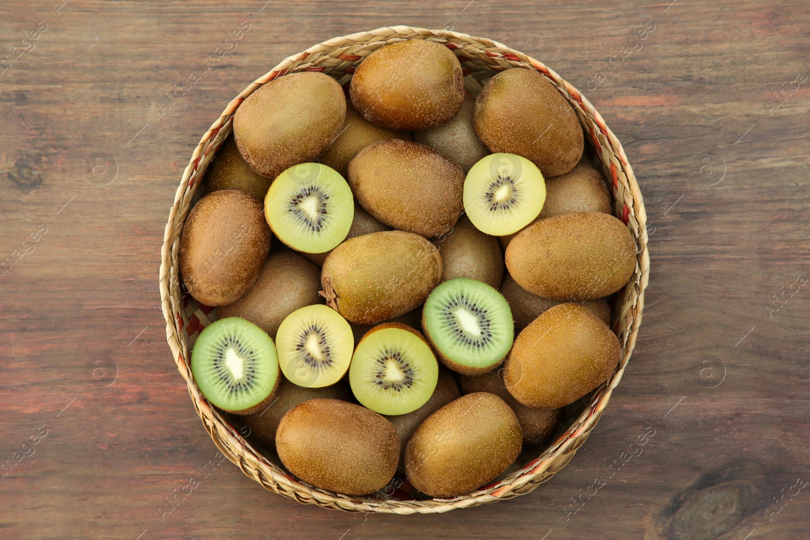 Photo of Basket of many whole and cut fresh kiwis on wooden table, top view