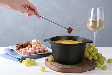 Woman dipping piece of bread into fondue pot with tasty melted cheese at white wooden table against gray background, closeup