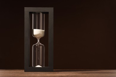 Photo of Hourglass with flowing sand on wooden table against dark brown background, space for text