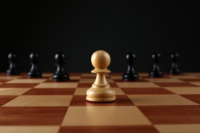 White pawn in front of black ones on wooden chess board against dark background