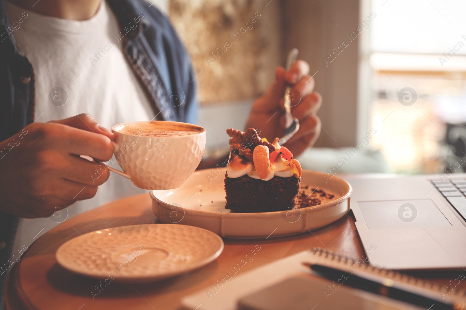Photo of Young blogger with laptop eating dessert and drinking coffee at table in cafe, closeup