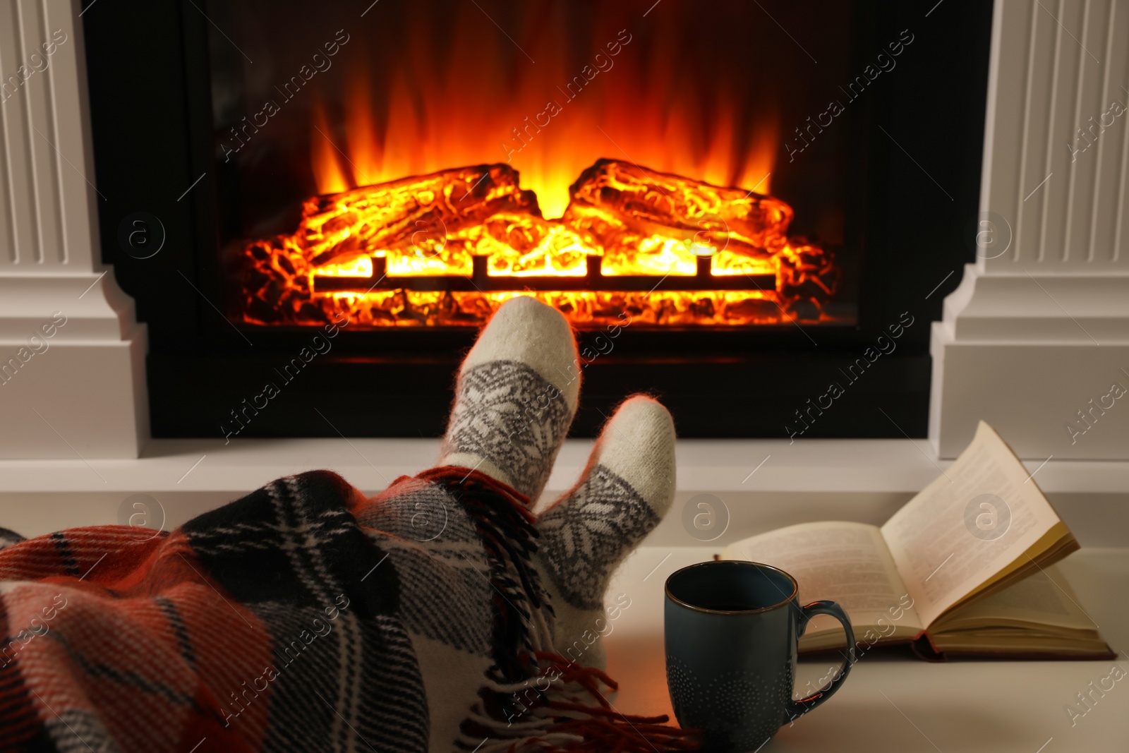 Photo of Woman with cup of hot drink and book resting near fireplace at home, closeup