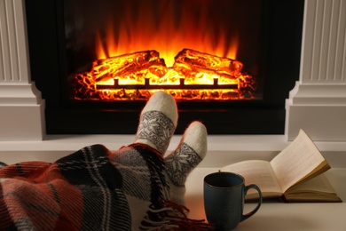 Photo of Woman with cup of hot drink and book resting near fireplace at home, closeup