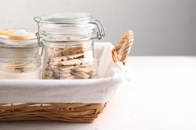 Photo of Glass jars with many wooden clothespins and laundry powder near candle in wicker basket on white table, space for text