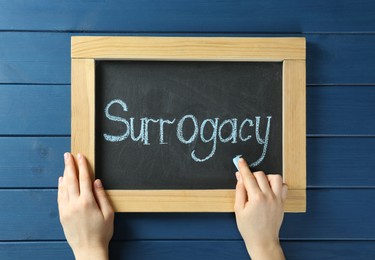 Photo of Woman writing word Surrogacy on small blackboard at blue wooden table, top view