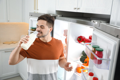 Young man drinking milk near open refrigerator in kitchen
