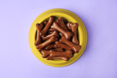 Yellow bowl with bone shaped dog cookies on purple background, top view