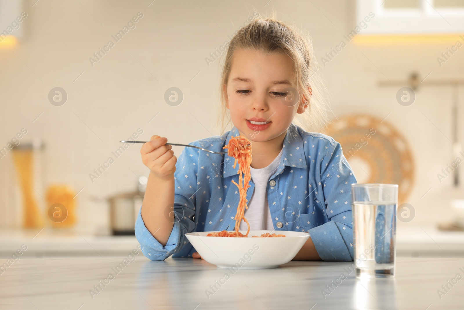 Photo of Cute little girl eating tasty pasta at table in kitchen