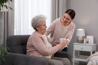 Photo of Young caregiver giving tea to senior woman in room. Home care service